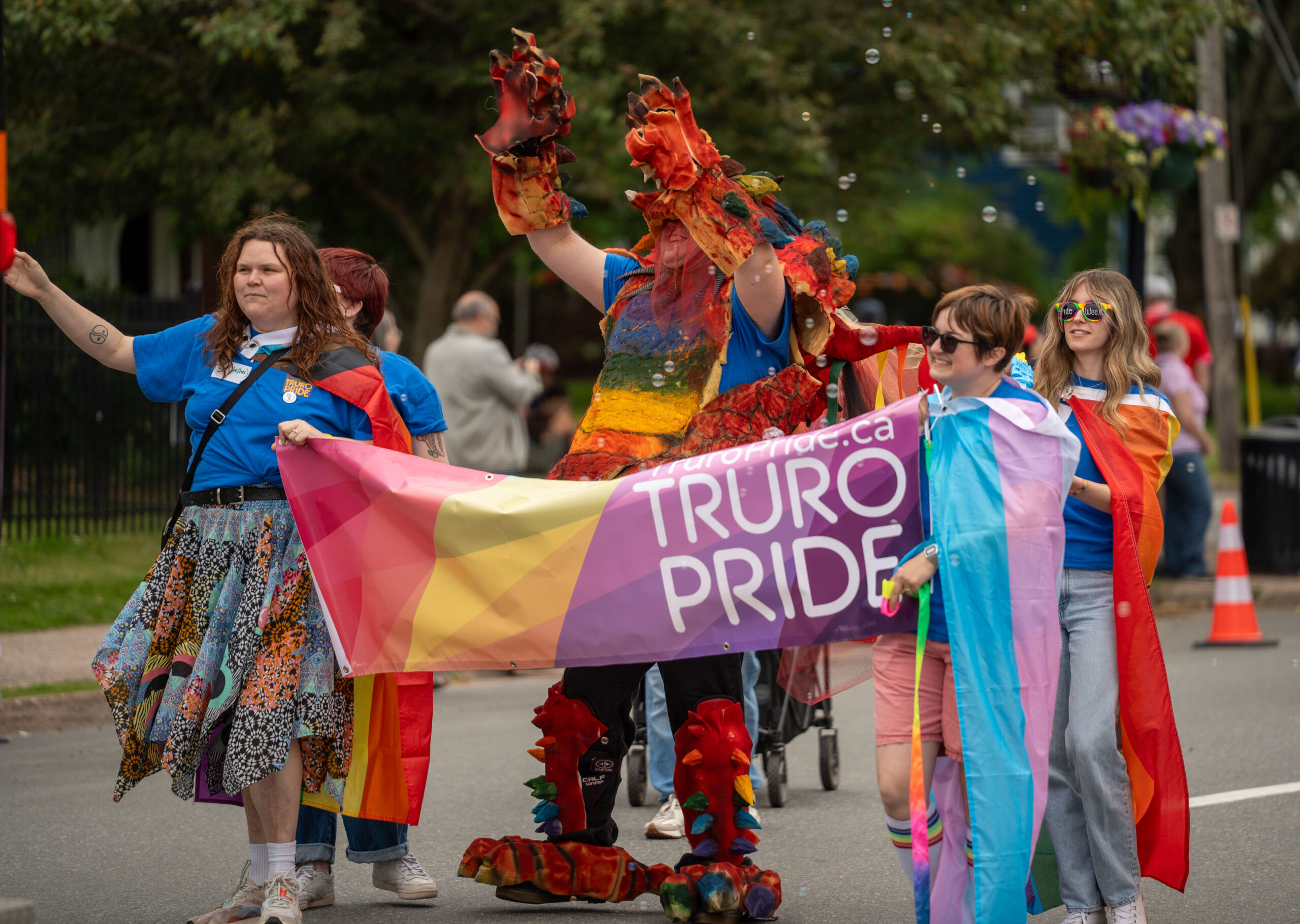 A photo from the 2025 Truro Pride Parade in downtown Truro. Two people wearing a pride flag and a trans flag carry rainbow coloured banner with Truro Pride printed on the front. Behind the banner is a large man in a dragon costume with his arms raised in the air in joyous celebration.